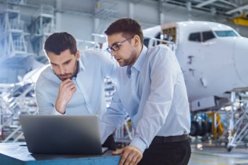 Zwei junge Männer schauen in ein Loptop, im Hintergrund eine Flugzeug-Montage